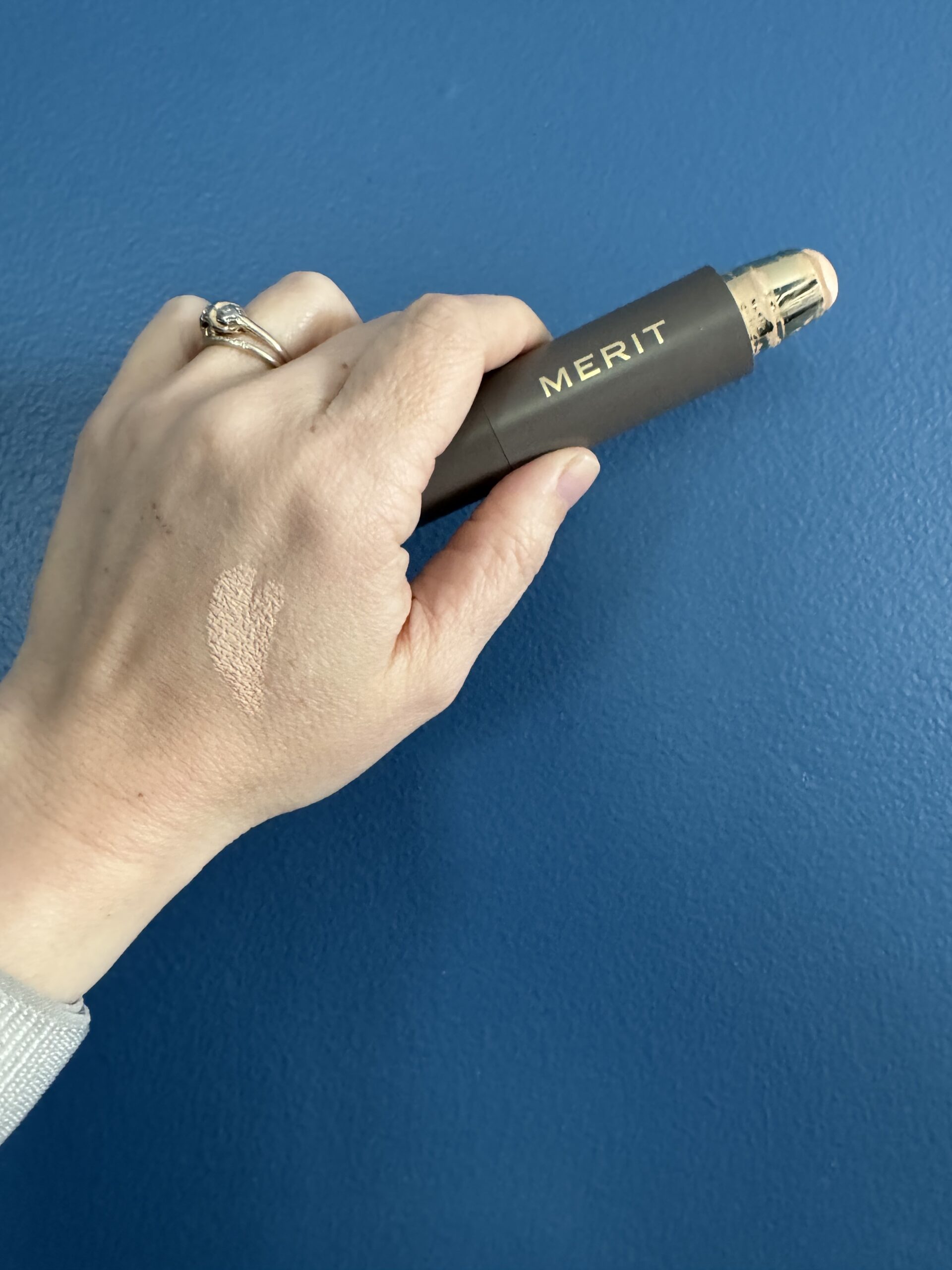 A hand against a blue background displaying swatched foundation makeup on the thumb and holding a tube of merit brand beauty product.