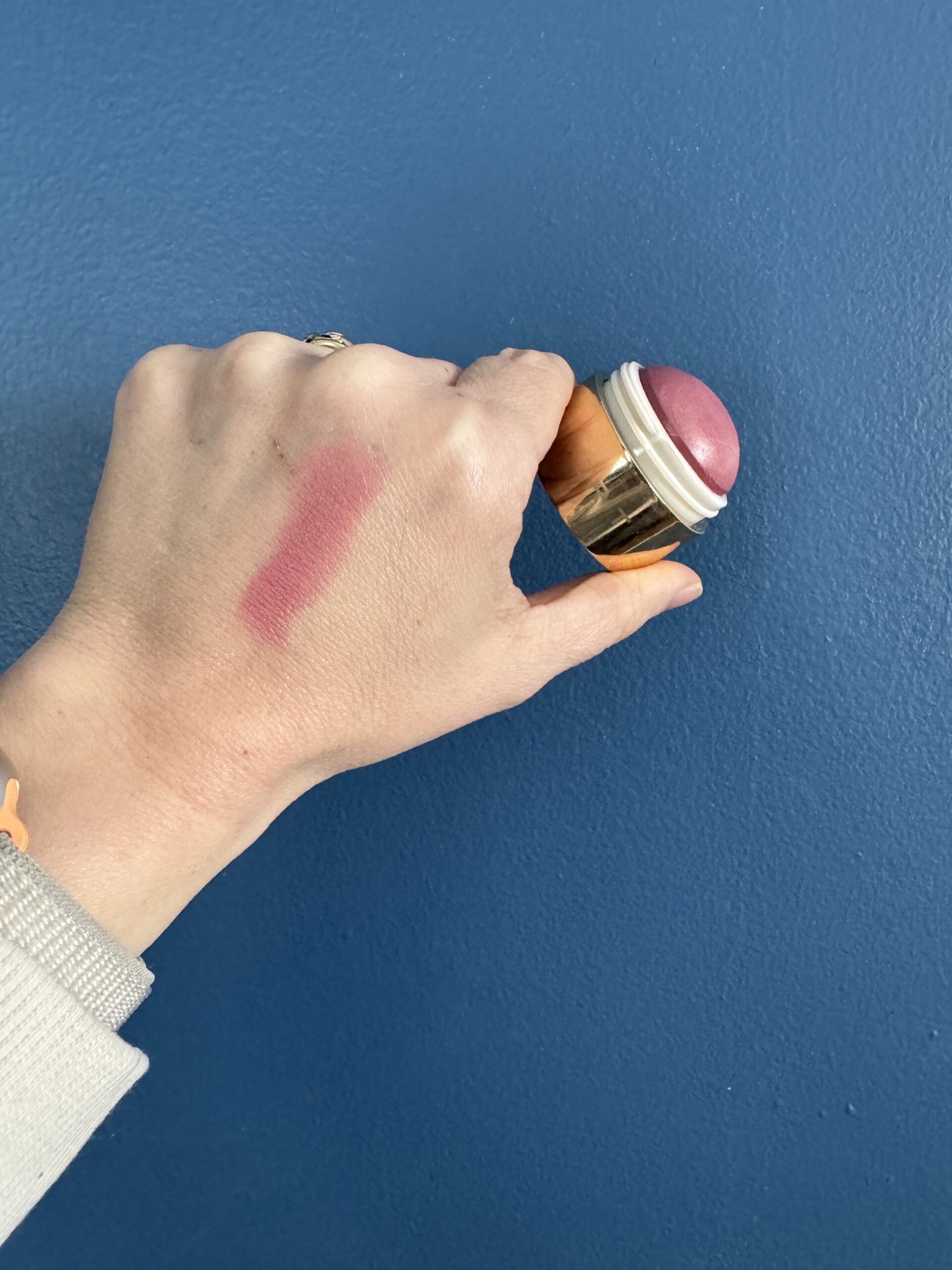 A hand holding a cosmetic product against a blue background with a swatch of pink makeup on the skin.
