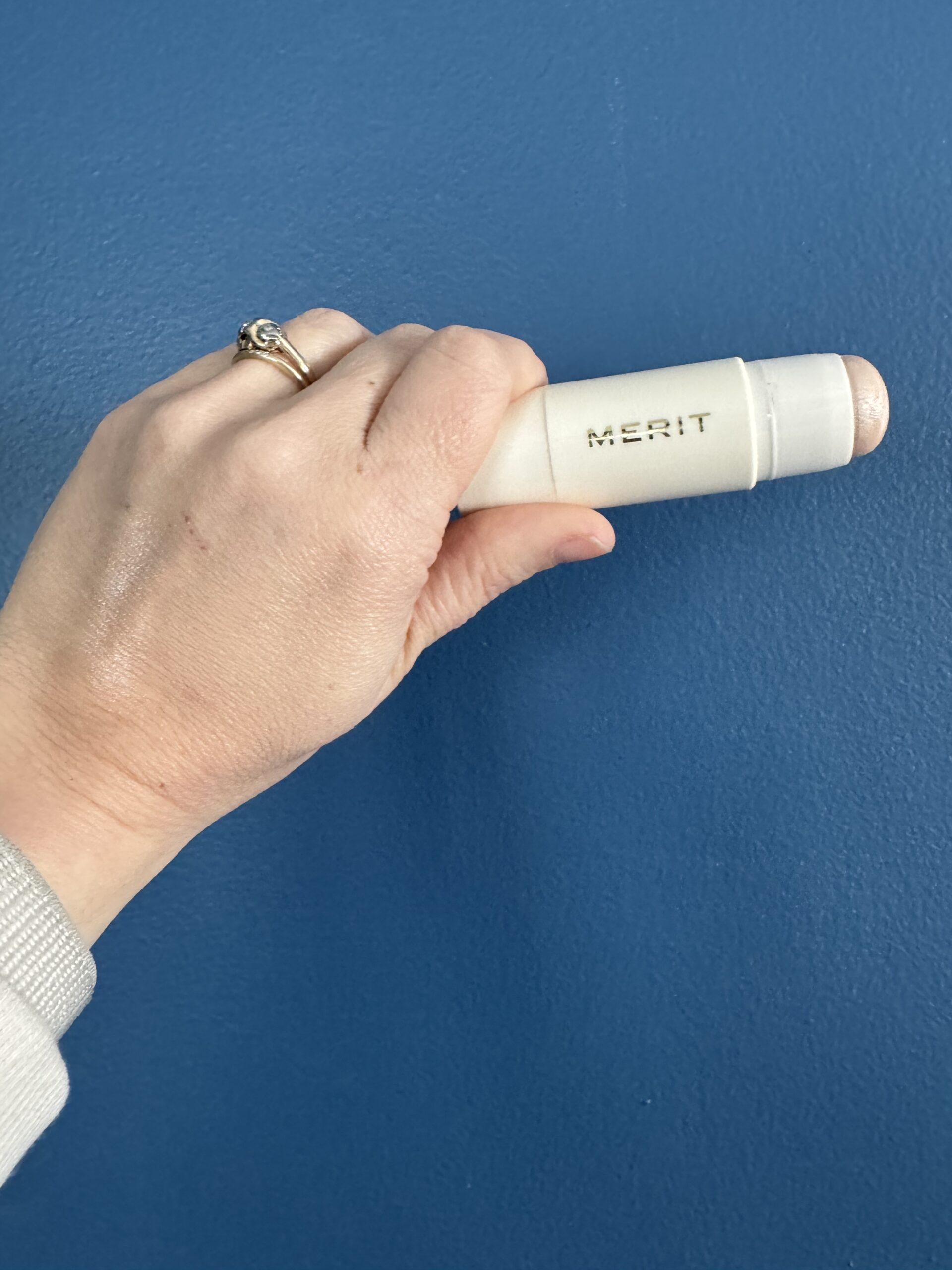 A hand holding a merit brand cosmetic product against a blue wall.