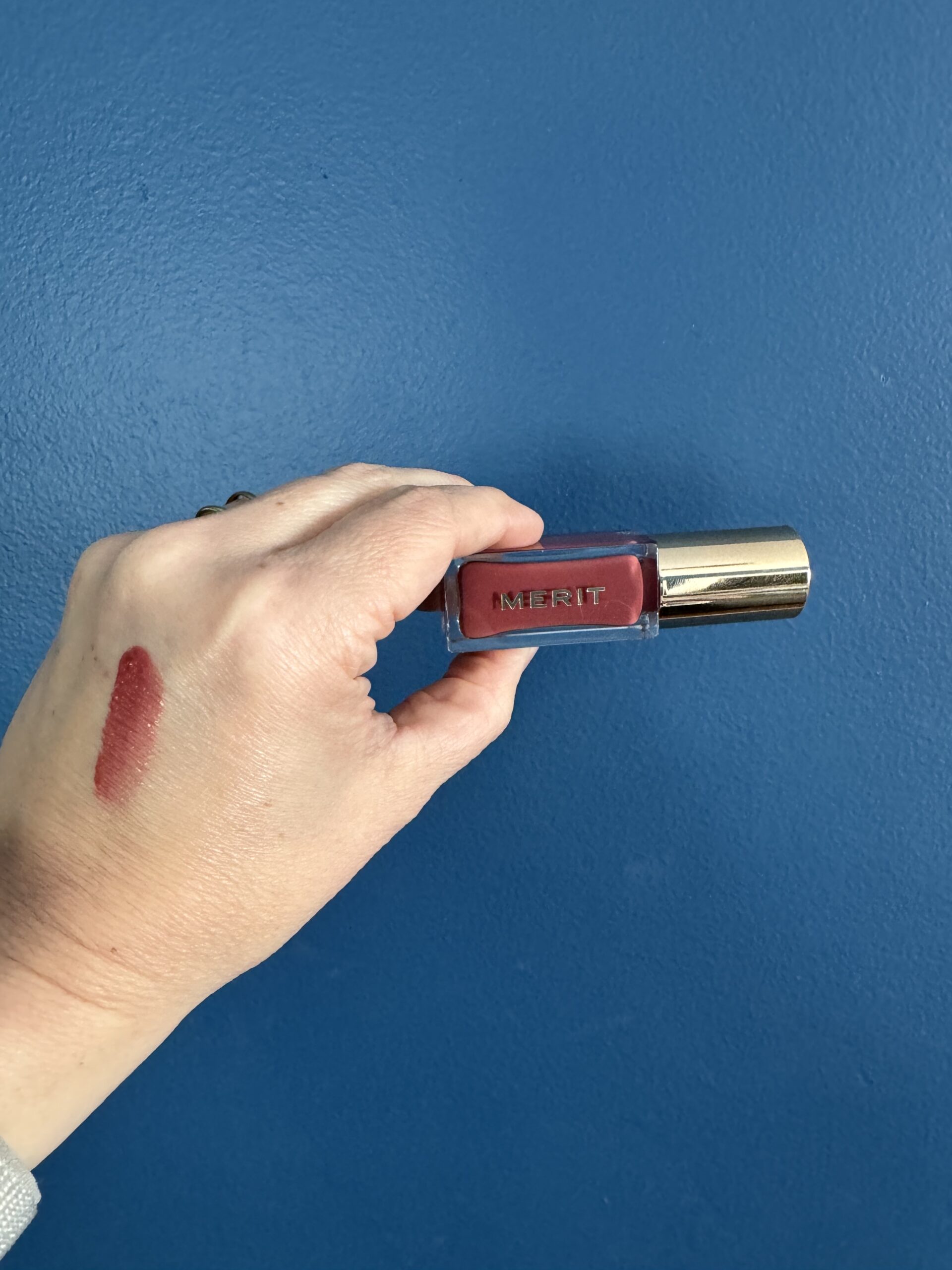 A hand holding a tube of merit lipstick against a blue background, with a swatch of the lipstick color on the person's wrist.