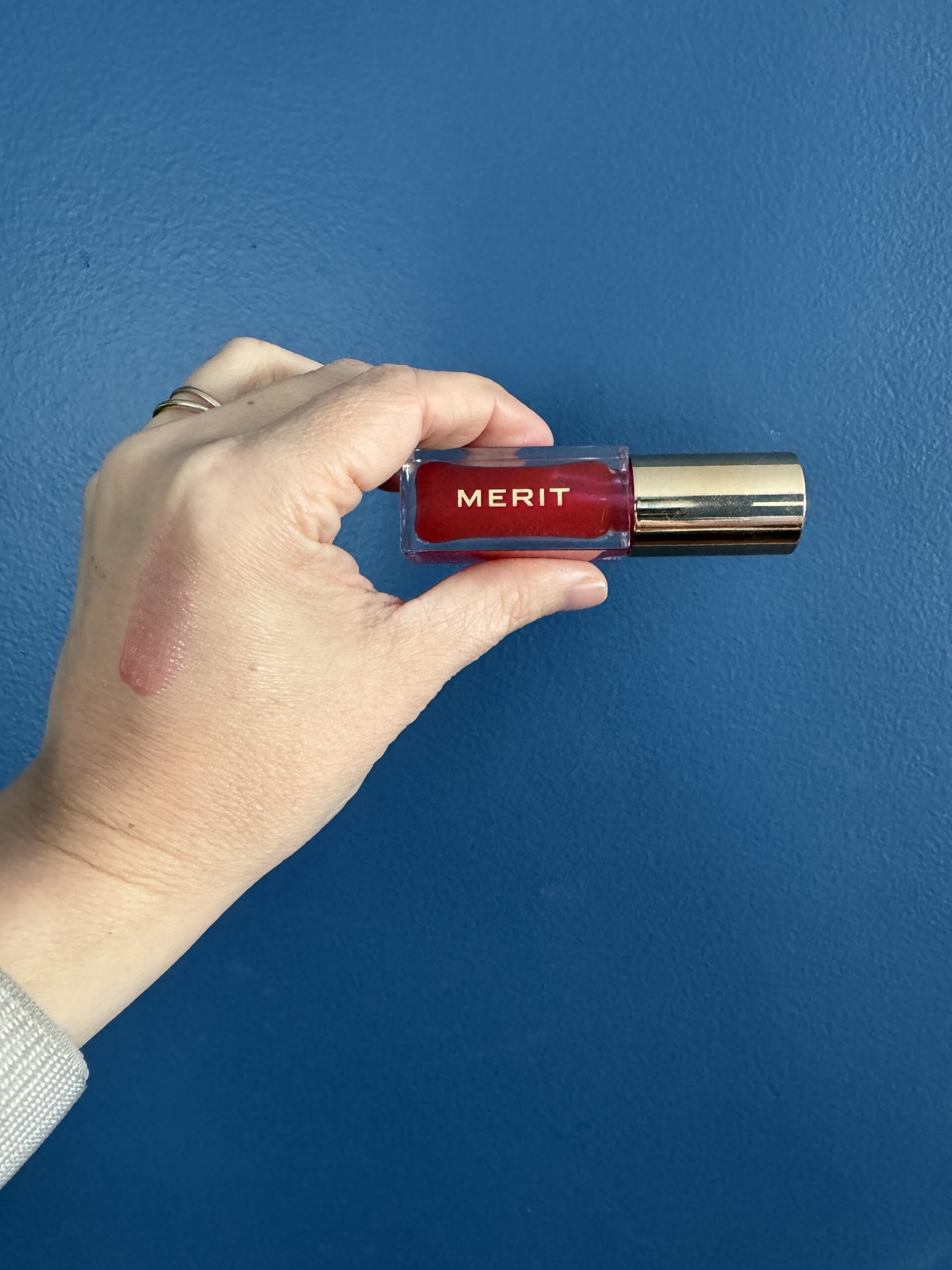 A person's hand against a blue background holding a tube of merit brand lip color with a swatch of the product on their wrist.