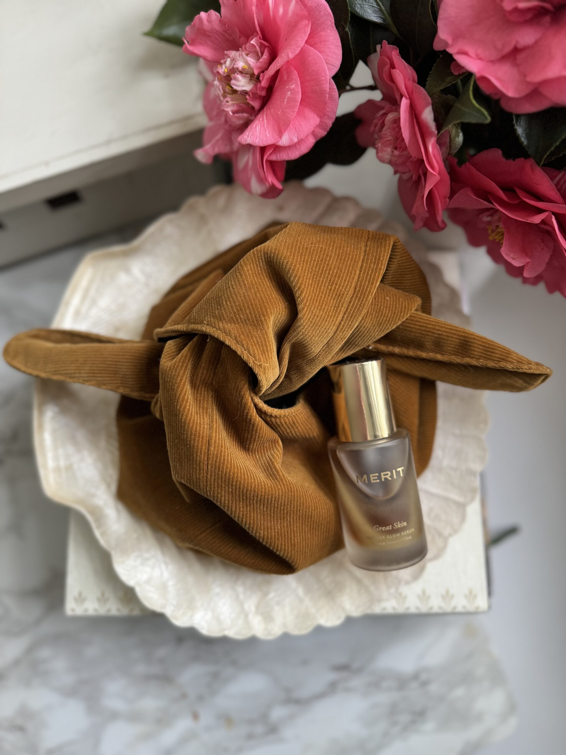 A beauty product placed on a textured brown cloth next to pink flowers on a white surface.