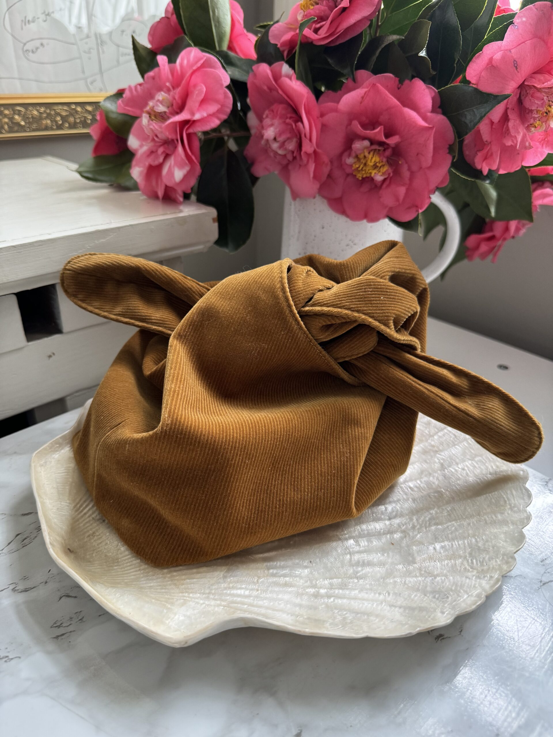 A mustard yellow fabric pouch with a knot, resting on a white decorative plate, next to pink flowers.