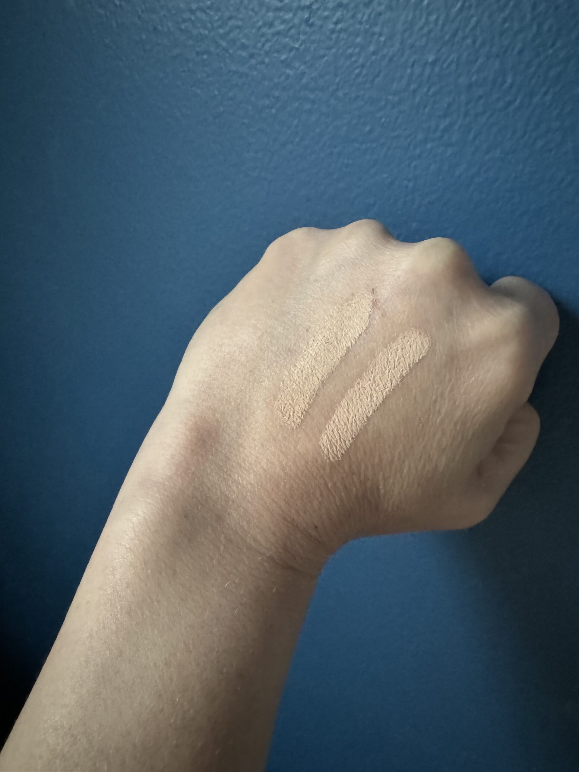 Swatches of two shades of foundation makeup applied to a wrist against a blue background.