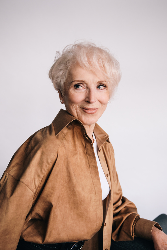 An elegant senior woman with short blonde hair, wearing a brown jacket and a white shirt, smiling gently at the camera against a light background.
