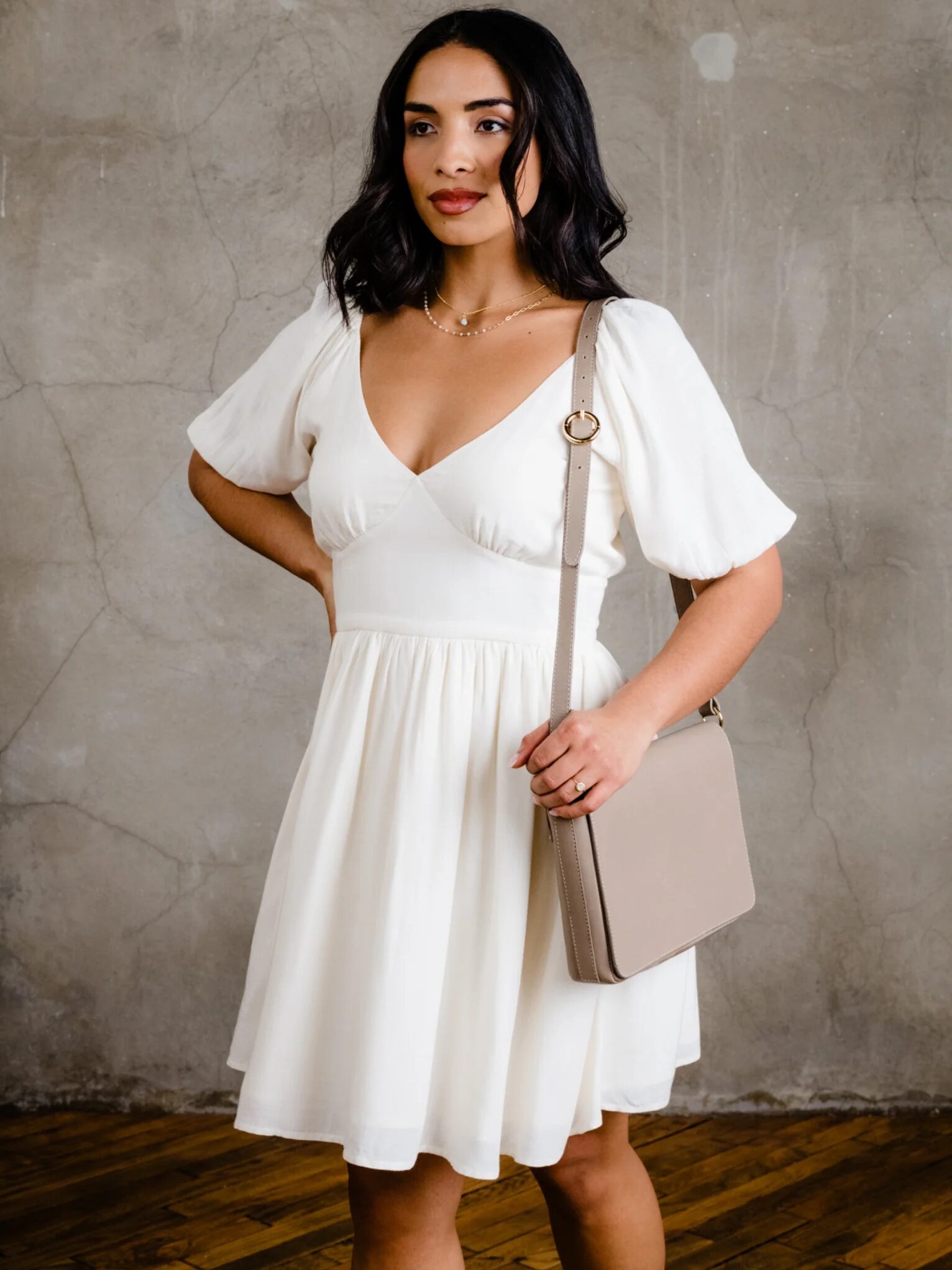 Woman in a white dress holding a beige shoulder bag standing against a textured wall.