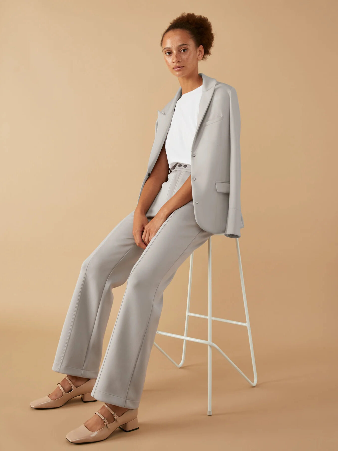 A woman is sitting on a stool in a grey suit.
