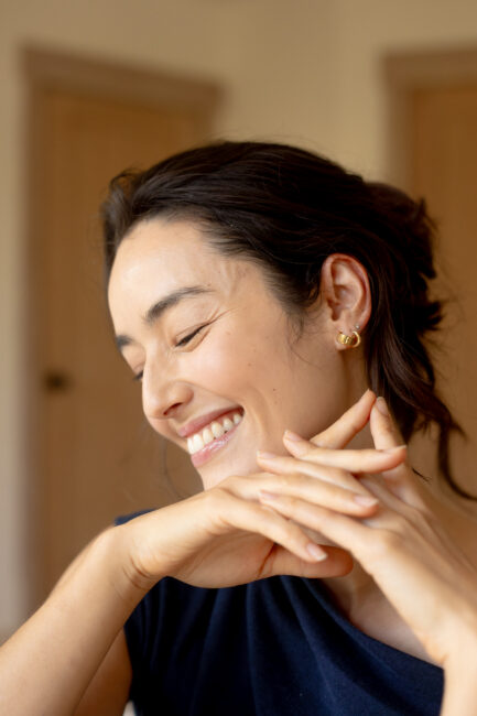 Woman with dark hair and gold earrings smiles with eyes closed, resting her chin on her hands. Neutral background with wooden doors.