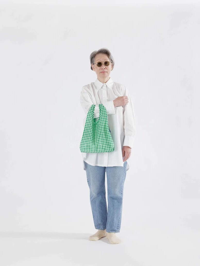 A woman standing against a white background, wearing sunglasses, a white blouse, blue jeans, and holding a green checkered tote bag.