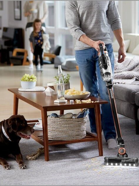 Person using a cordless vacuum cleaner in a living room while a dog watches and another person is in the background.