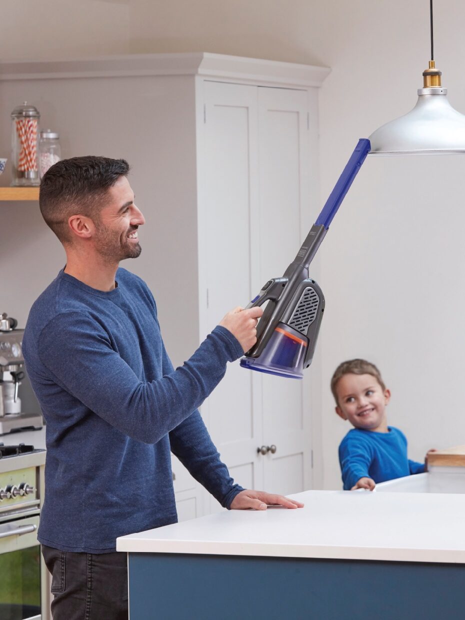 A man uses a cordless vacuum cleaner in the kitchen as a child watches from behind the counter.