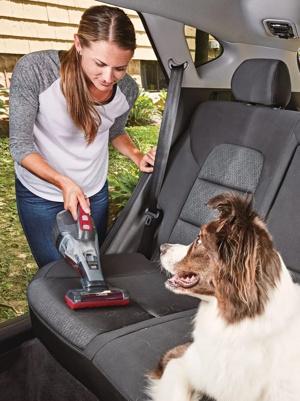 Woman using a handheld vacuum to clean the backseat of a car while a dog watches.