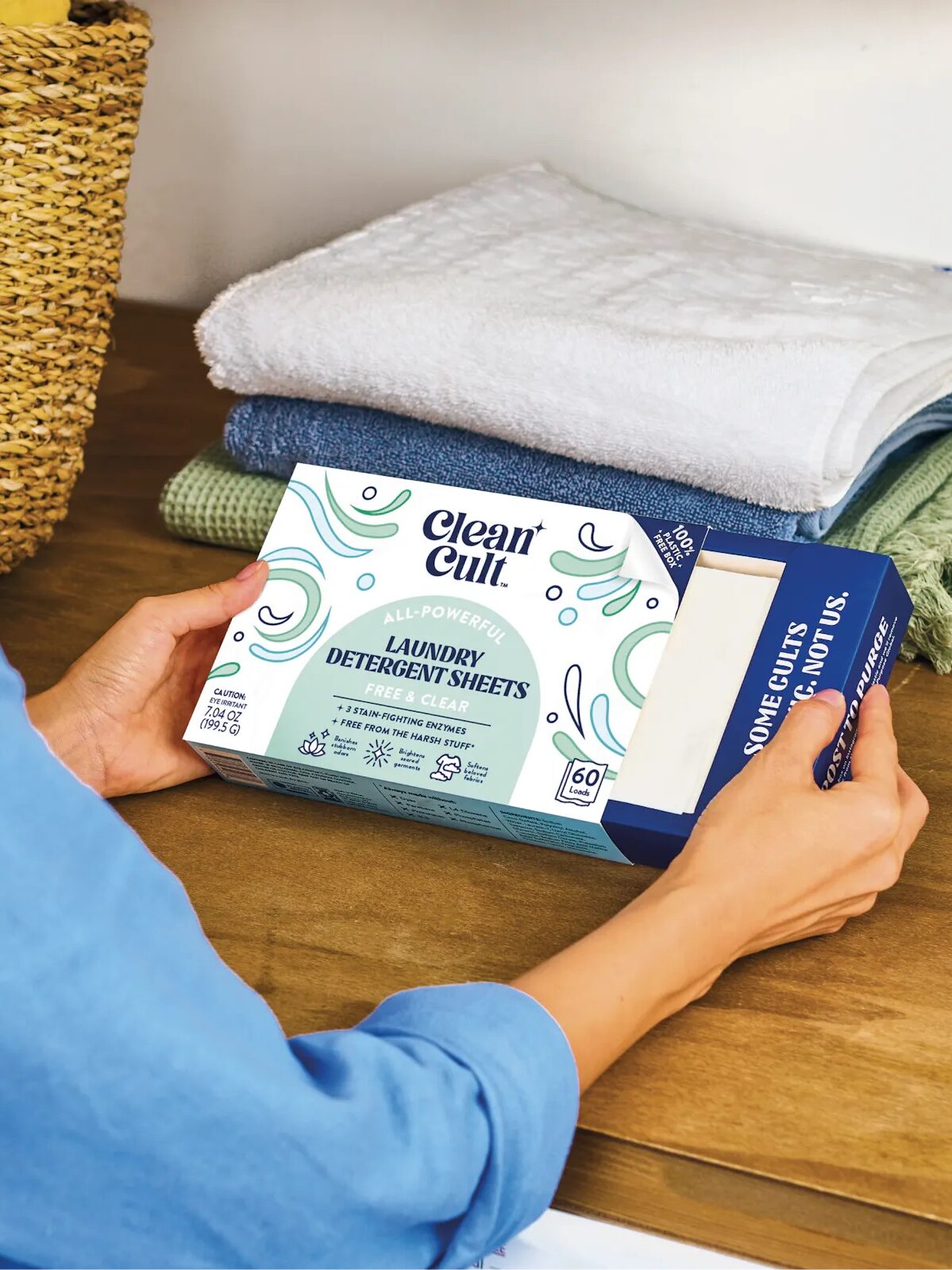 A person holding a box of cleancult laundry detergent sheets next to a pile of folded towels.