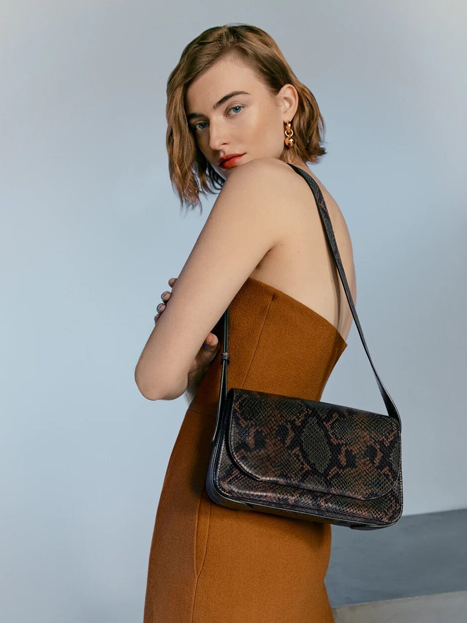 A woman in a brown dress glances over her shoulder, showcasing a shoulder bag with a snakeskin pattern.