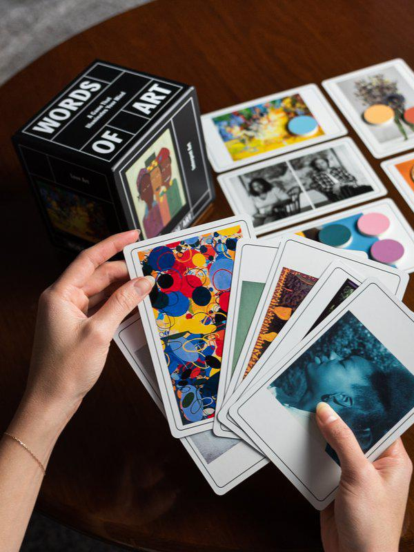 Hands holding a deck of art-themed playing cards with more cards spread out on a wooden table.