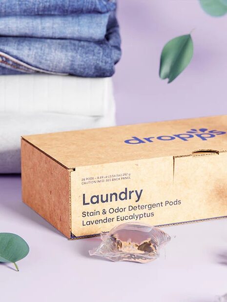 A box of dropps laundry detergent pods with a lavender eucalyptus scent, displayed with folded clothes and lavender sprigs on a purple background.