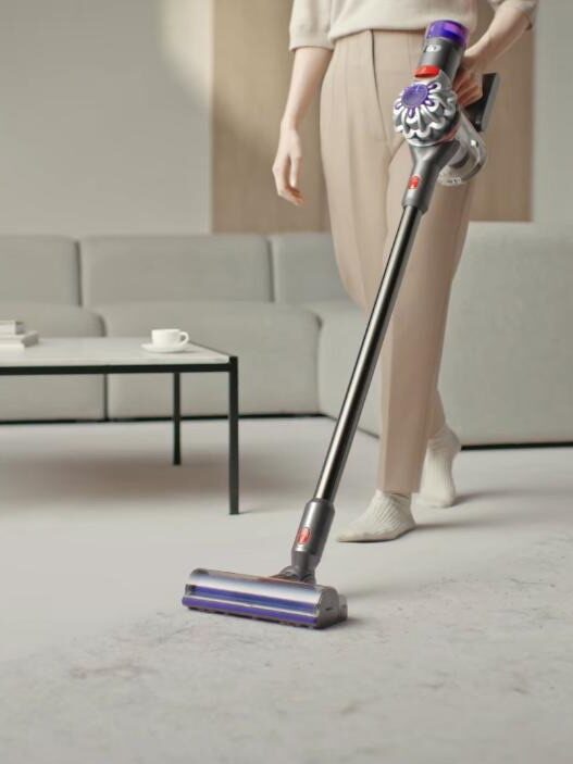 A person using a cordless stick vacuum cleaner on a light-colored floor in a modern living room setting.