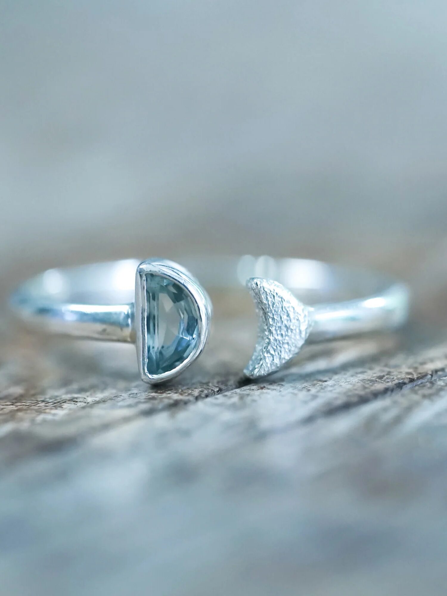 A silver ring with a moon-shaped split design, featuring one smooth and one textured half, resting on a wooden surface.
