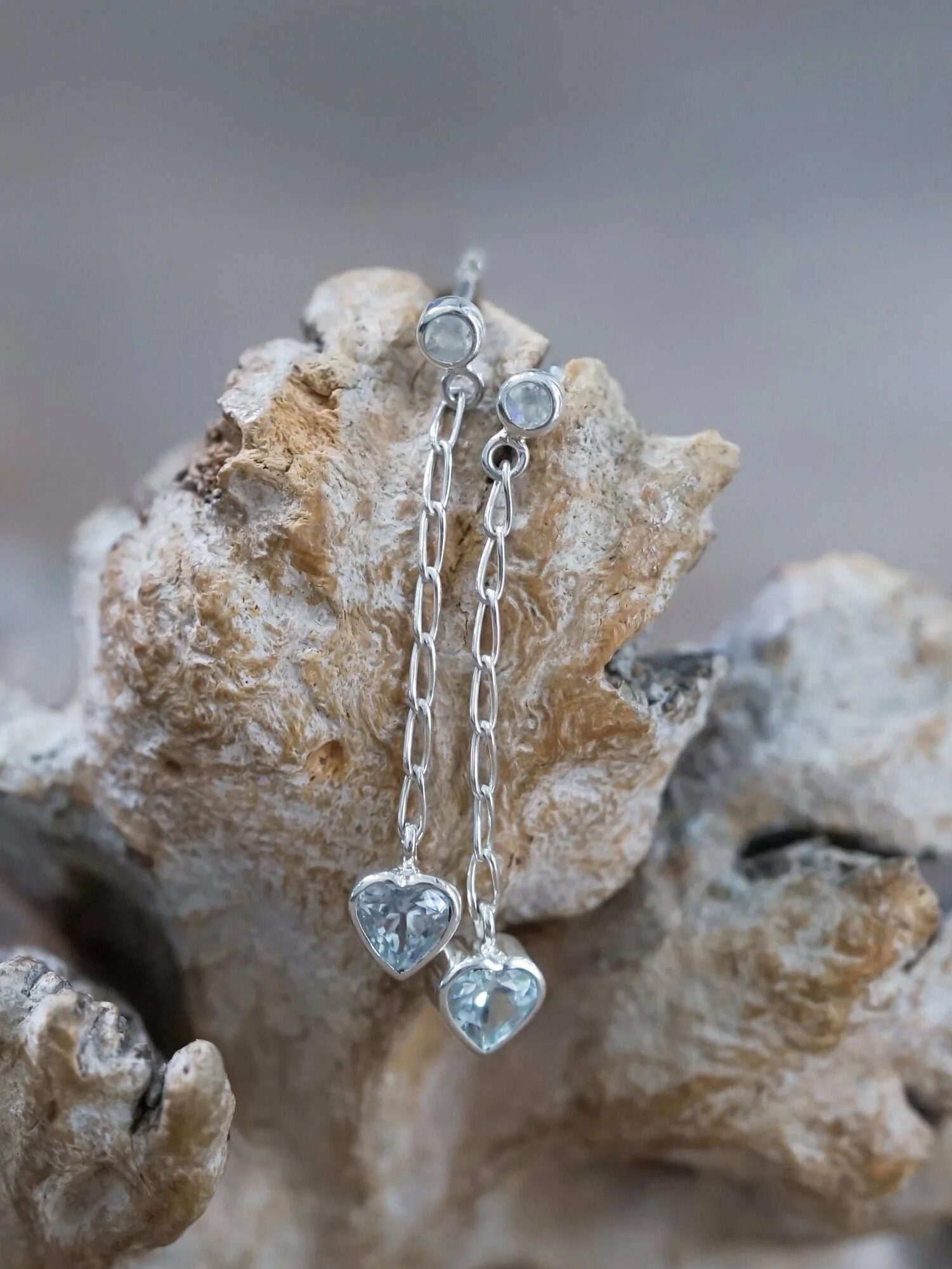 A pair of heart-shaped pendant earrings displayed on a piece of driftwood.