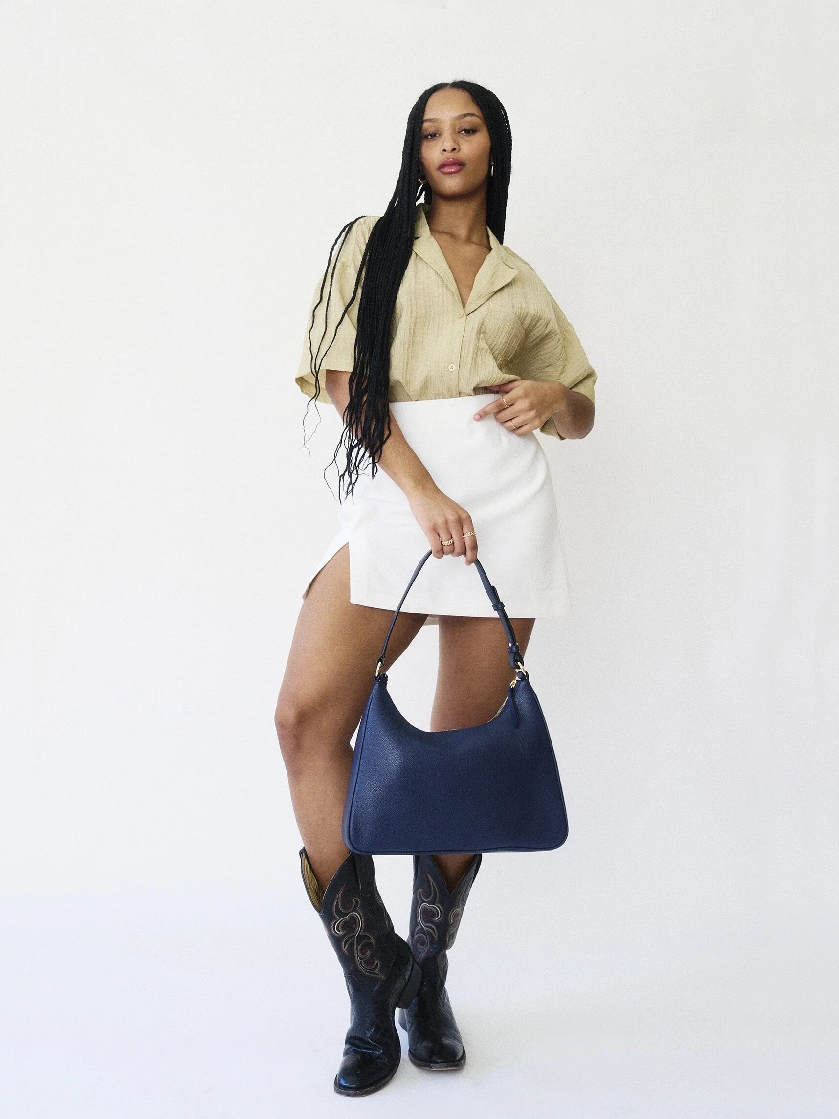 A woman posing with a blue handbag, wearing a white skirt, beige blouse, and black boots.