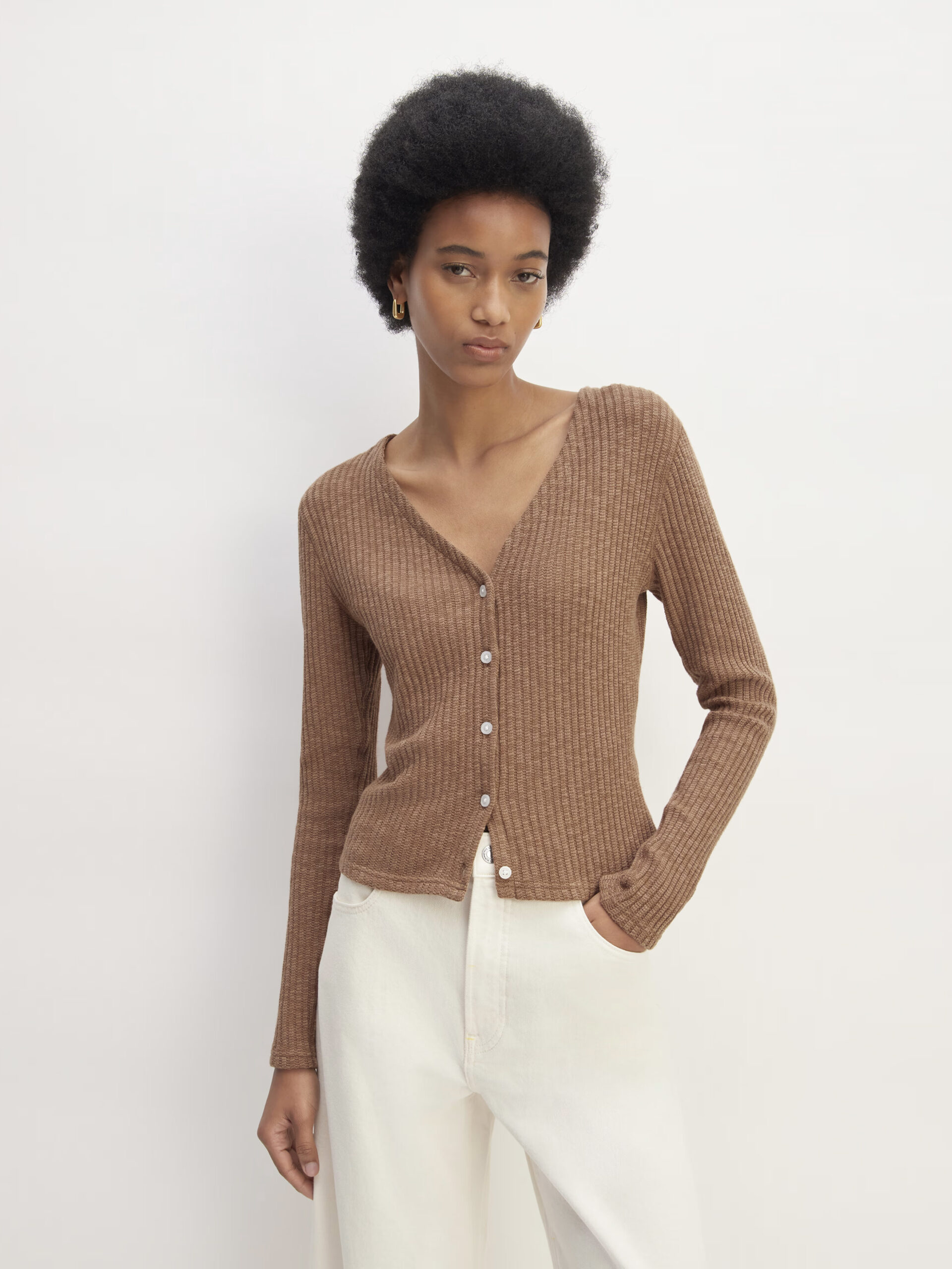 Woman in a brown cardigan and white pants posing against a plain background.
