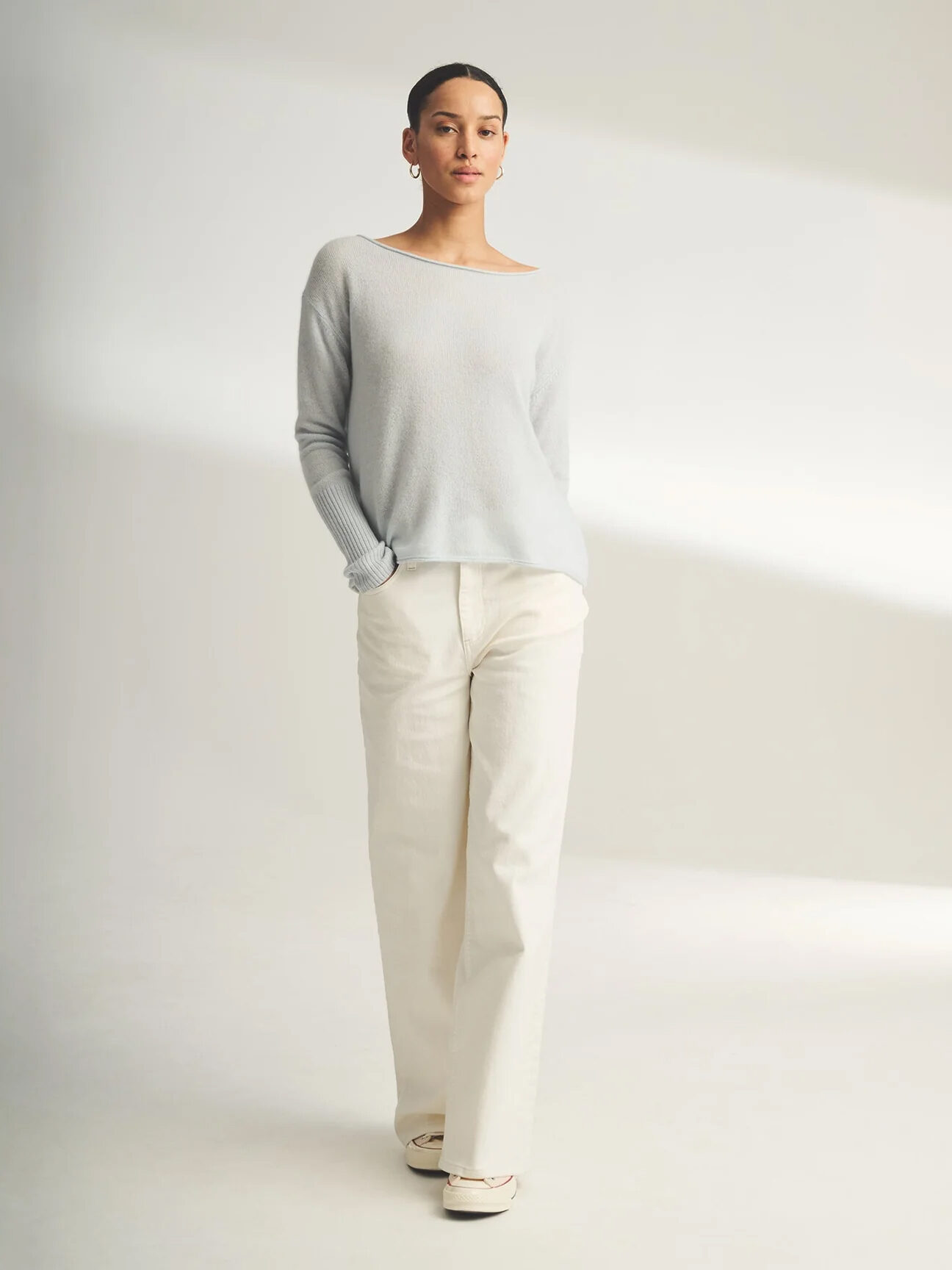 Woman in a light grey sweater and white trousers standing against a neutral background.