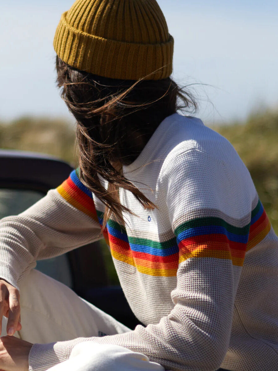 Person in a white sweater with rainbow stripes sitting outdoors with hair blowing in the wind.