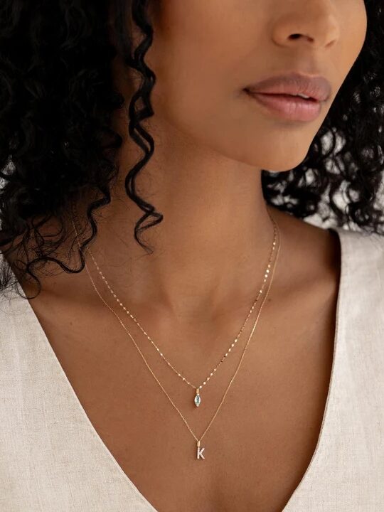 A woman wearing a layered necklace with a pendant and a personalized initial charm.