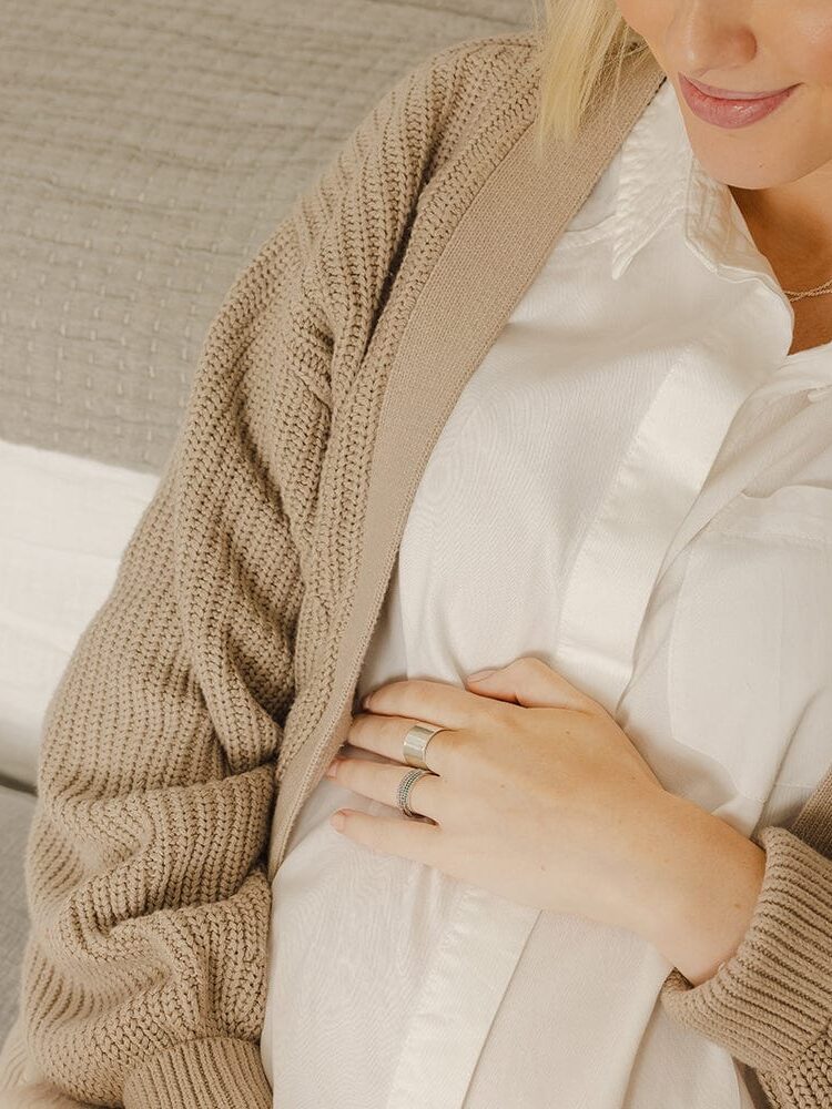A smiling woman gently holding her pregnant belly while sitting on a couch, wearing aquamarine rings