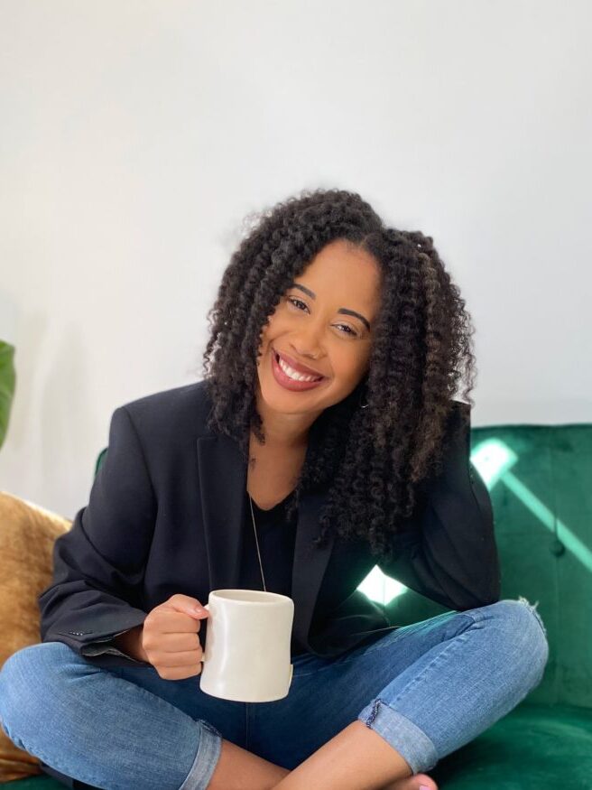 A therapist smiles at the camera, seated on a couch and holding a cup of coffee.  