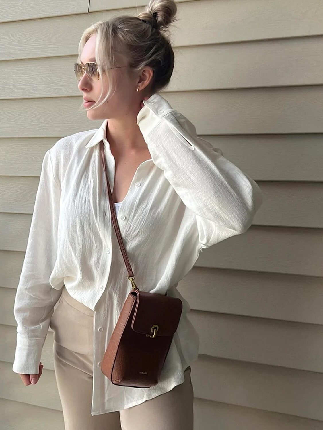 Woman in a white shirt and sunglasses posing with a small brown shoulder bag.