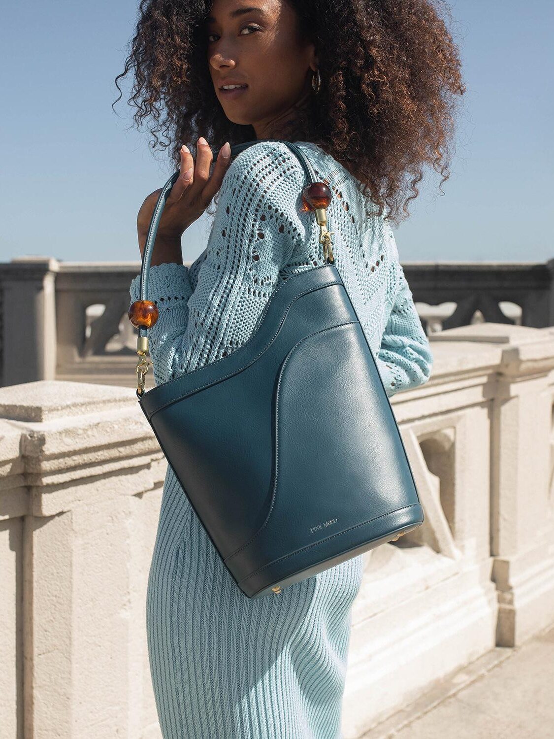 Woman posing with a stylish leather handbag on a sunny balcony.