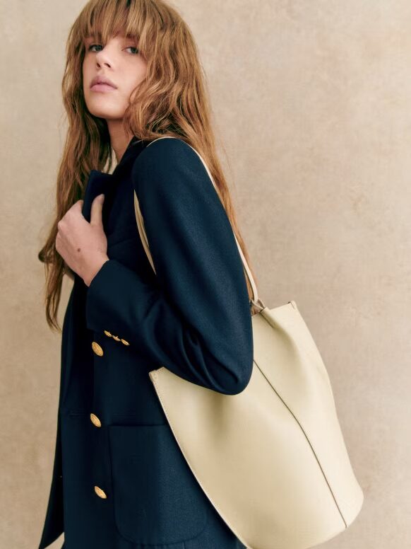 Woman with bangs carrying a large beige tote bag and wearing a dark coat with gold buttons.