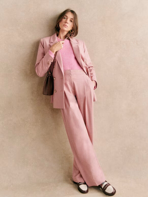 A woman in a pink suit and pink sweater leaning against a wall.