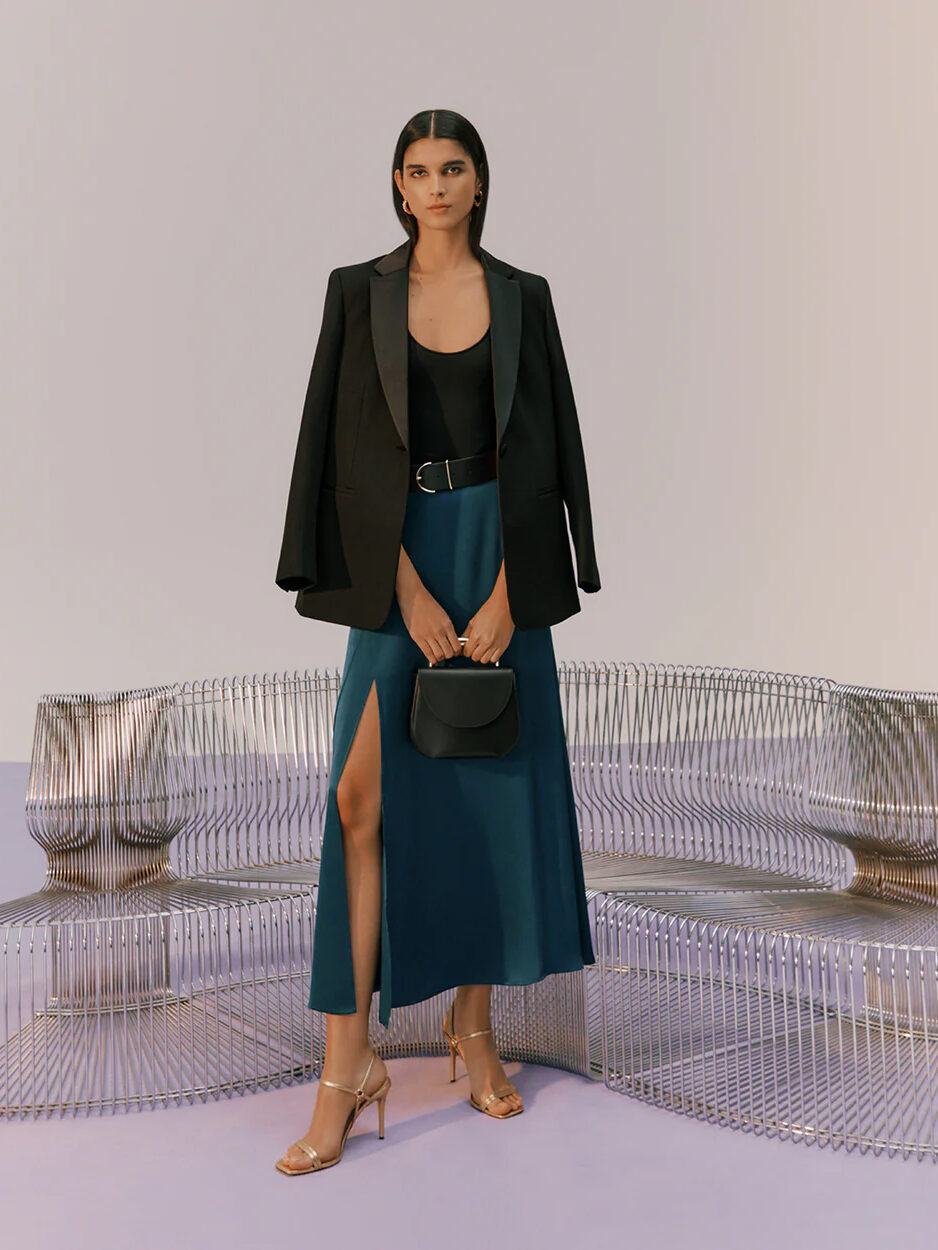 A woman stands wearing a black blazer over a sleeveless top, paired with a high-slit blue skirt, accessorized with a belt, a small bag, and strappy heels.