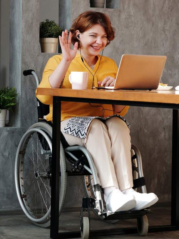 An individuals seated in a wheelchair, waving to a therapist via their laptop. 