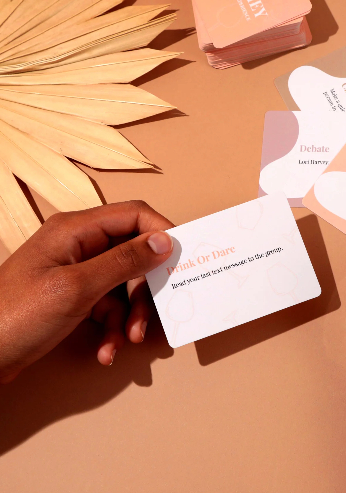 A hand holding a "drink or dare" card with the dare "read your last message to the group" against a backdrop of similar cards and palm leaves.