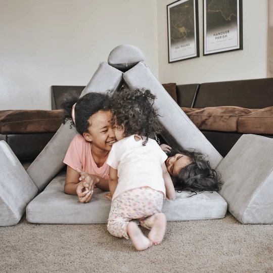 Two children giggling while playing with couch cushions arranged as a fort.
