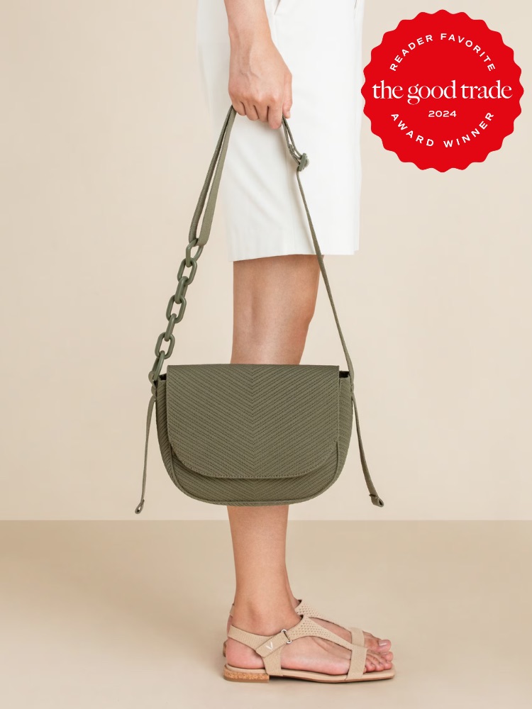 A person showcasing a fashionable green shoulder bag with a herringbone pattern, along with a white skirt and pink sandals against a neutral background. the image includes a badge indicating the product is a reader favorite from the good trade, awarded in 2024.
