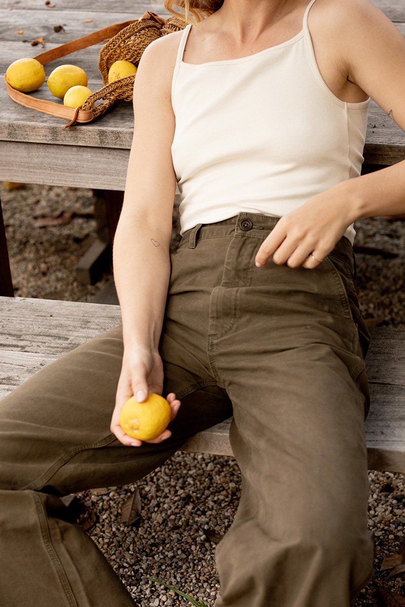 A person in a white tank top and olive green pants sitting on a wooden bench, holding a lemon, with more lemons nearby.