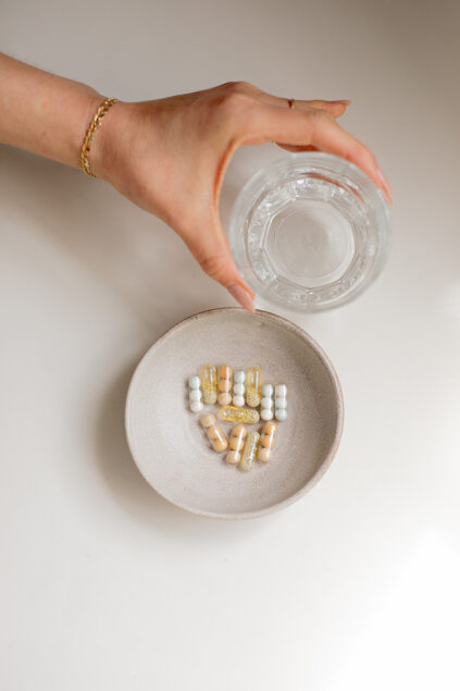 A hand pouring water from a glass into a ceramic bowl containing assorted prenatal vitamins and tablets arranged in a circle.