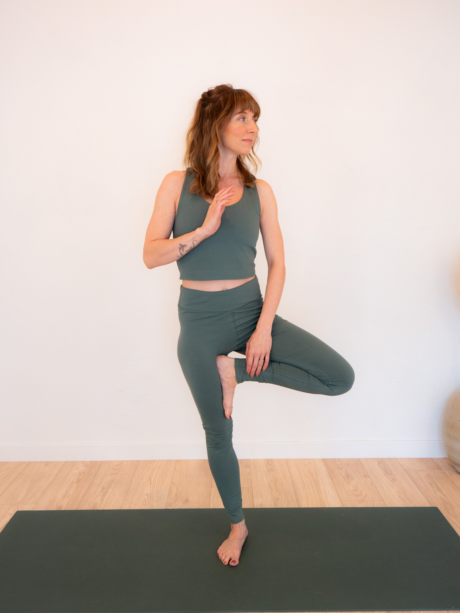 A woman in a green yoga outfit performs a tree pose on a yoga mat against a plain white wall.