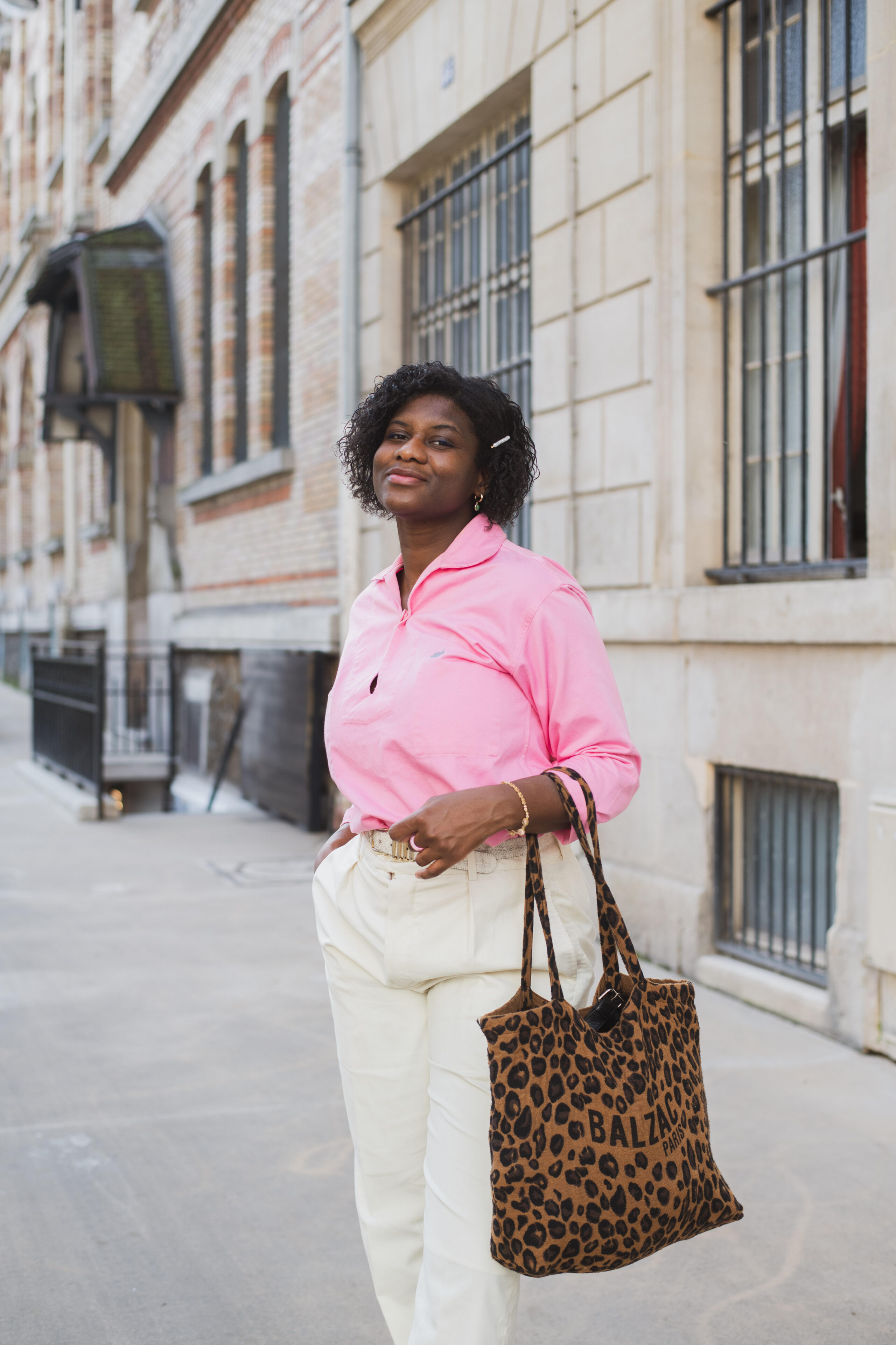 A woman in a pink shirt and white pants holding a leopard print bag stands confidently on a city street.