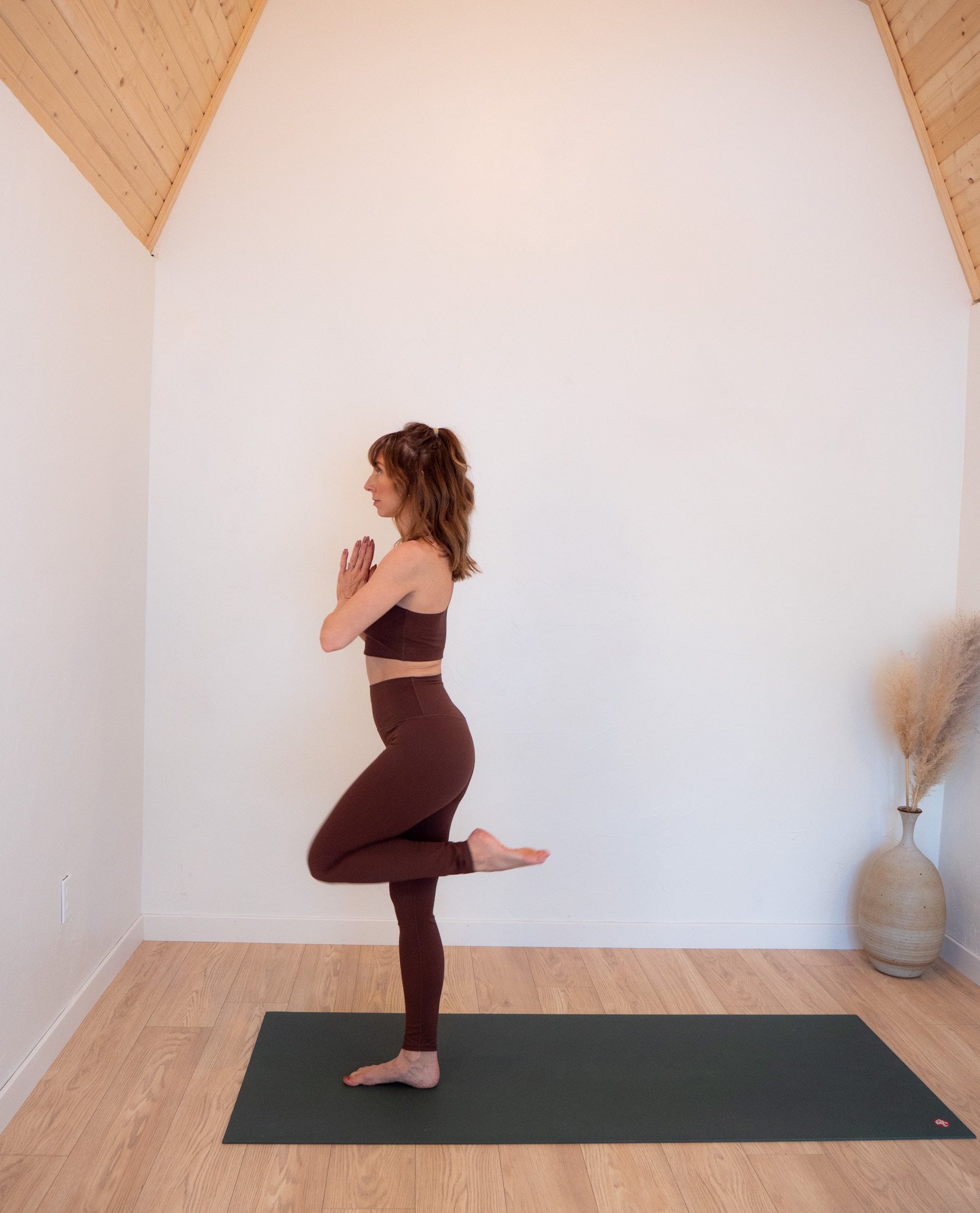 A woman in a brown yoga outfit performs a tree pose on a green mat in a serene, minimally decorated room with a slanted ceiling.