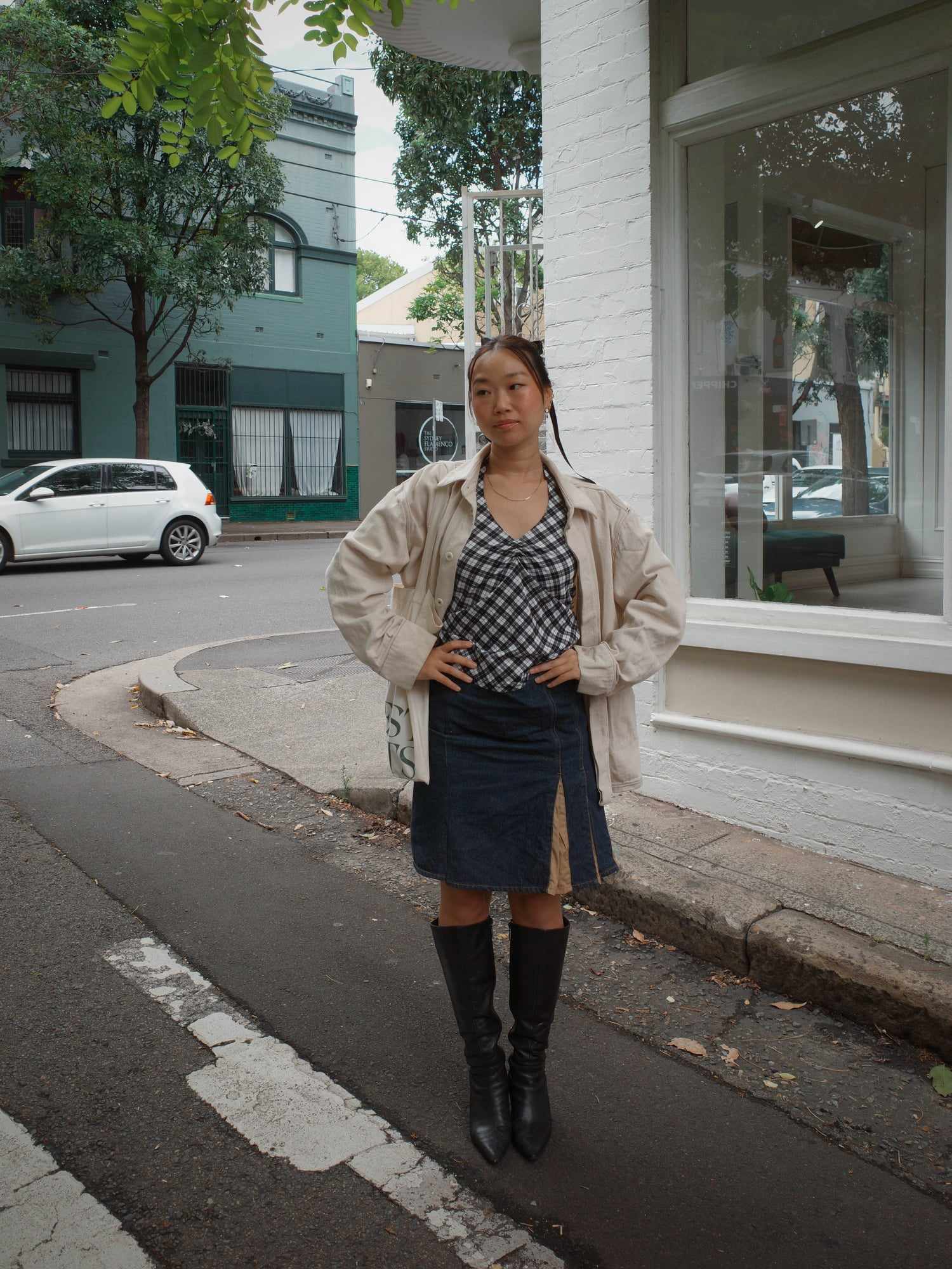 A woman stands on a street corner, wearing a trench coat, checkered blouse, denim skirt, and tall black boots, with buildings and a car in the background.