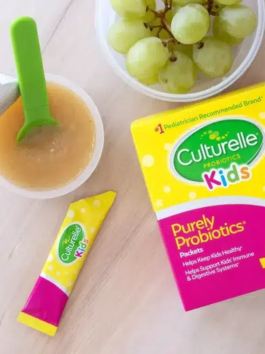 A box of culturelle kids probiotics alongside a bowl of applesauce and grapes, with a probiotic packet partially emptied into the applesauce.