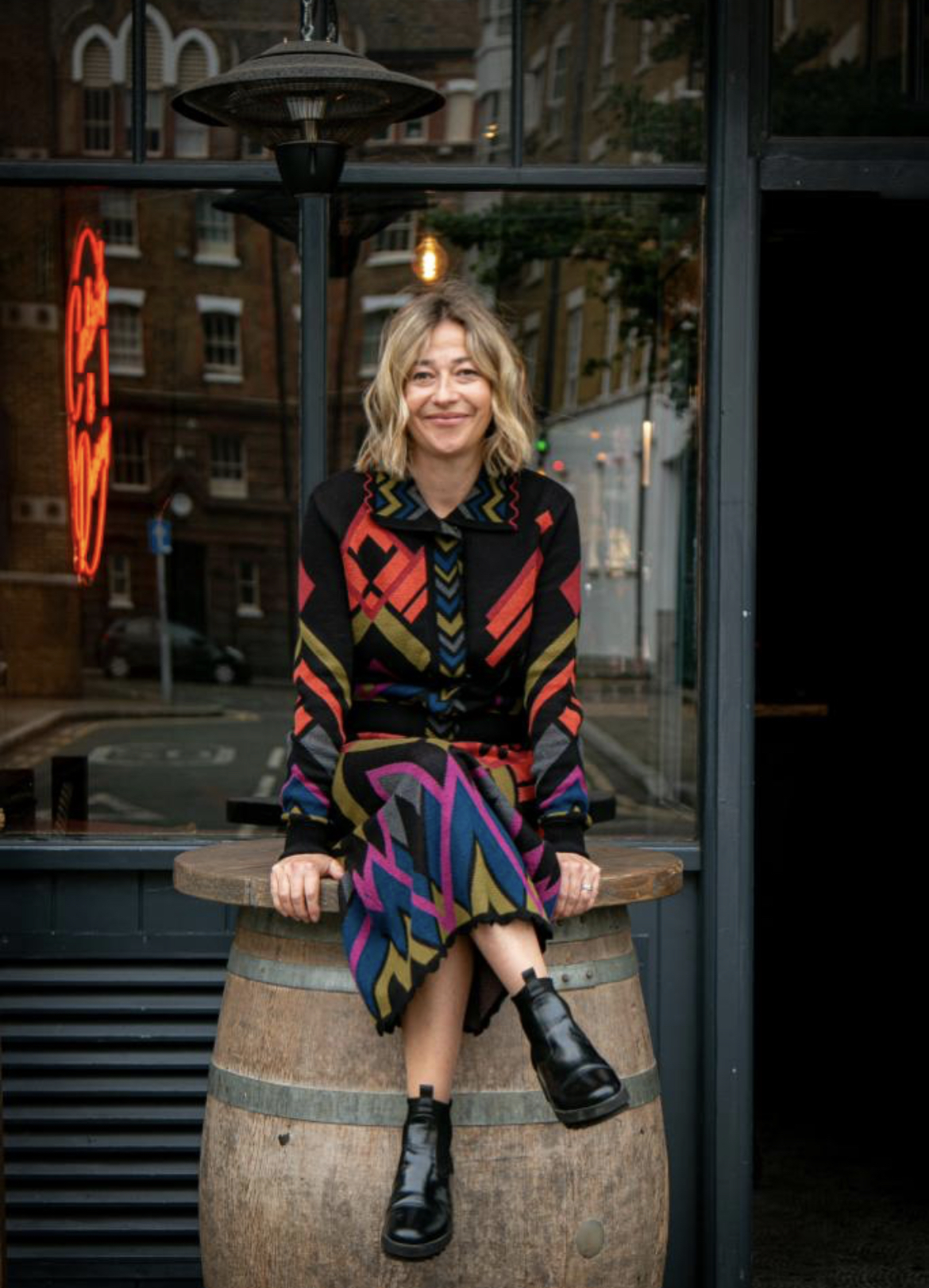 A woman in a colorful patterned outfit sits on a wooden barrel outside a cafe, smiling, with a neon sign visible through the window behind her.