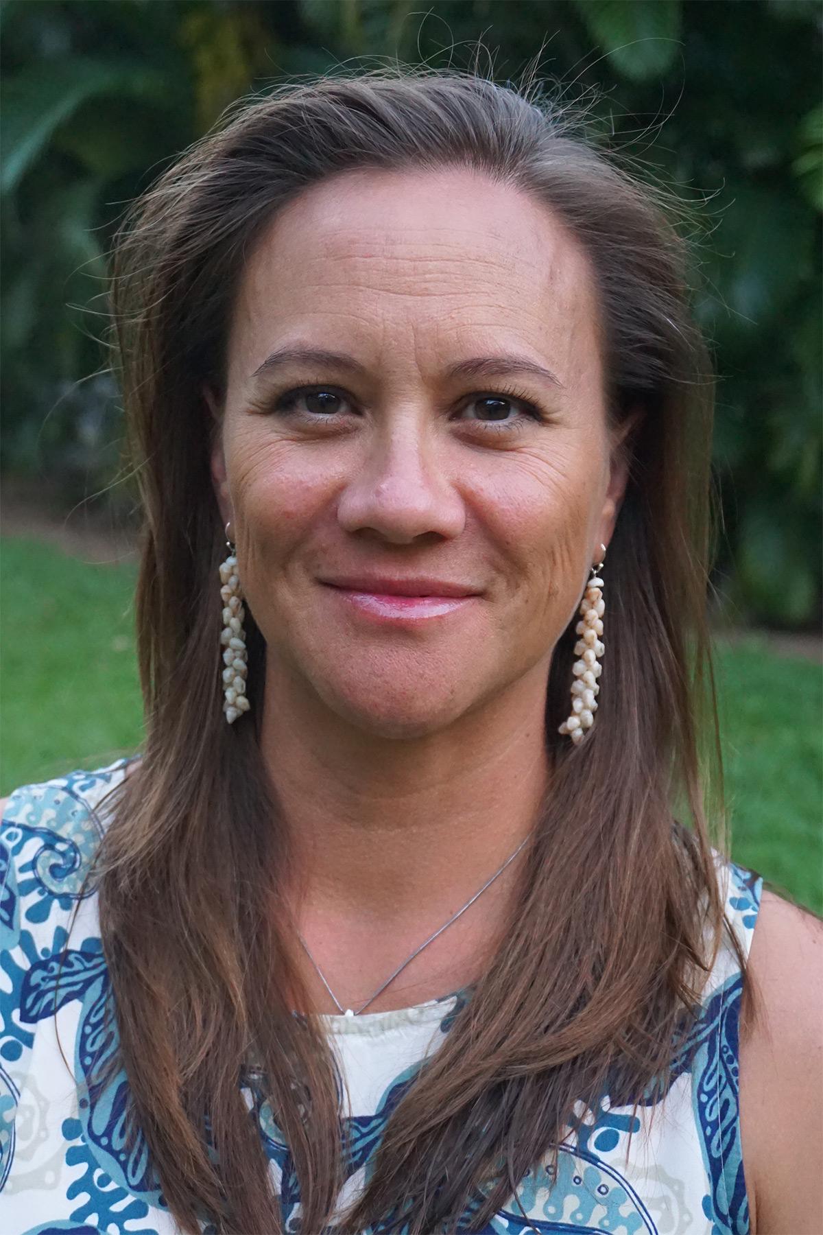 Portrait of a smiling middle-aged woman with shoulder-length brown hair, wearing large earrings and a floral top, standing outdoors.