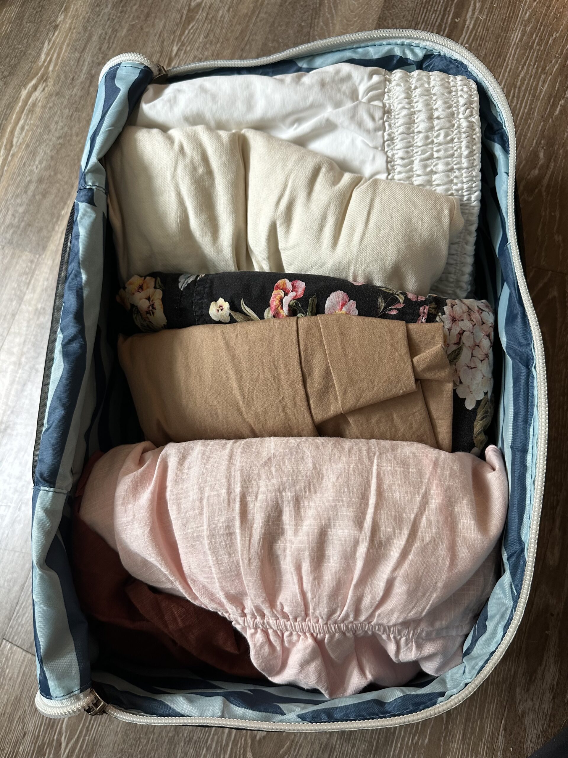 Open suitcase packed with folded clothes, including floral patterns and plain textiles, viewed from above on a wooden floor.