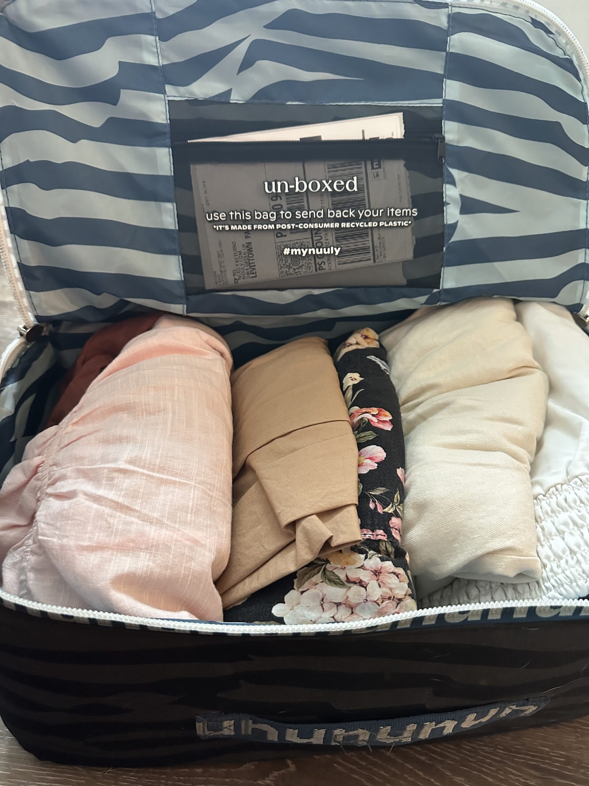 A suitcase open on a wooden floor, packed with neatly folded clothes in various colors and patterns, featuring a return shipping label inside the lid.